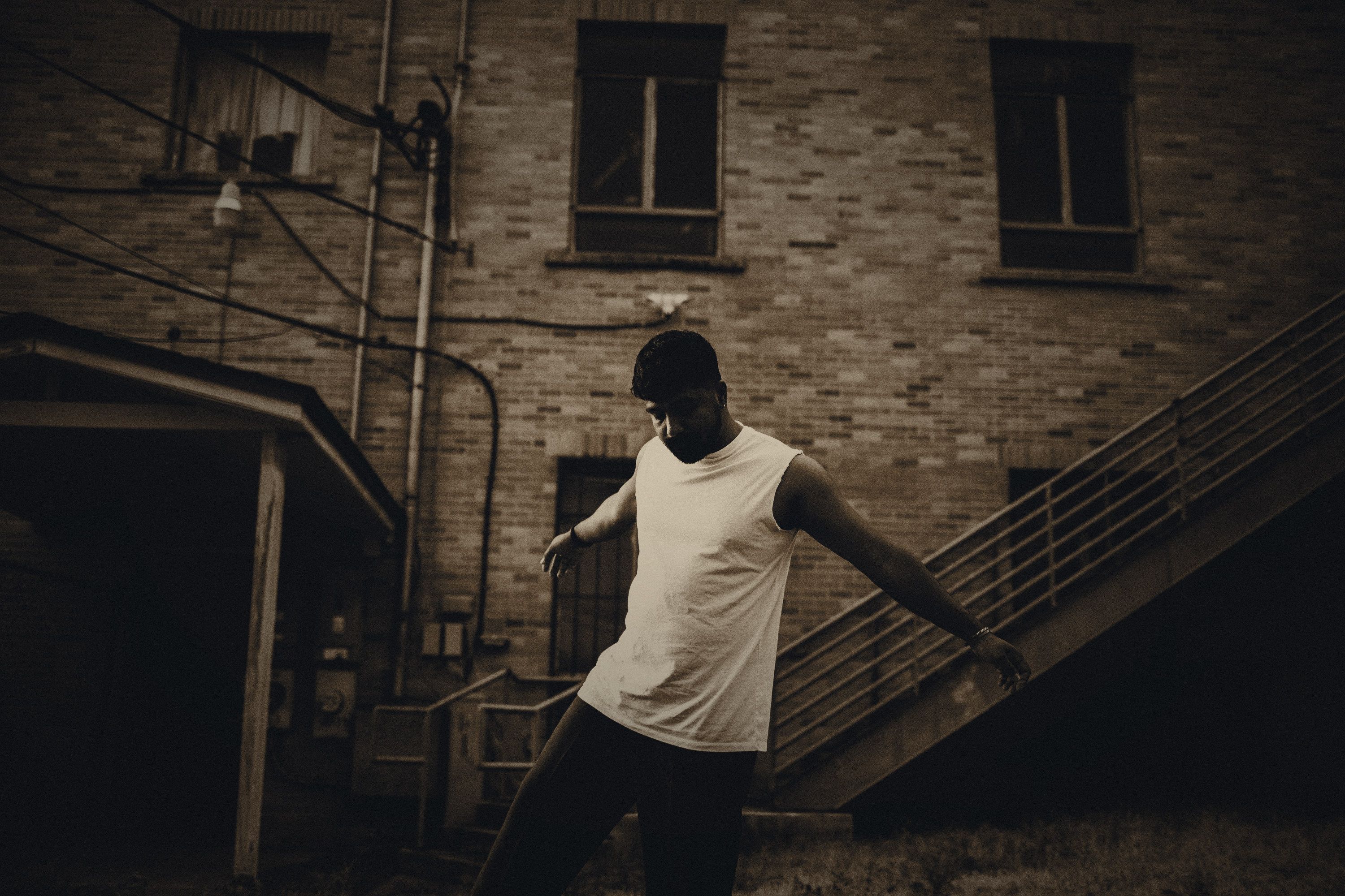 Adi Rao in a brick back lot at dusk, arms out and eyes down.